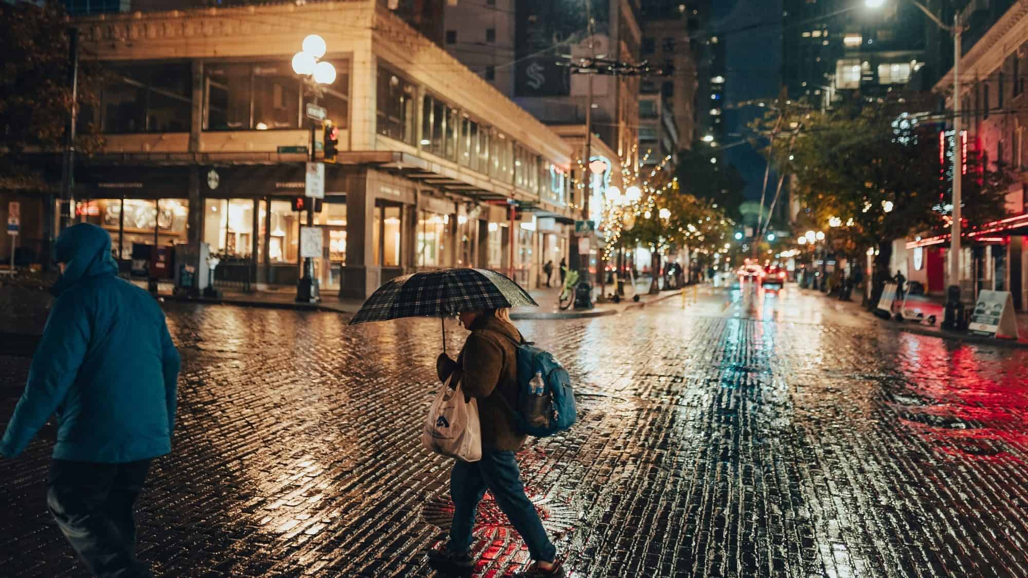 A person clutching an umbrella braves the soggy streets at night, navigating a city alive with lighted storefronts and illuminated streetlights, feeling the pulse of urban life.