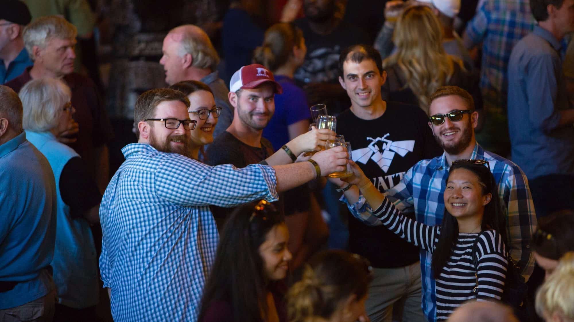 In a bustling Seattle scene, six people stand together, smiling warmly and raising glasses in a toast amidst the lively crowd. The crisp winter air adds a festive touch to their celebration.