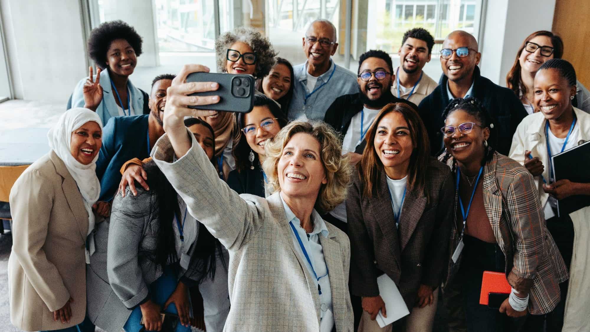 A diverse group of people in business attire, celebrating Women's History Month, pose for a selfie. They're all smiles, standing together in an indoor Seattle setting, radiating unity and empowerment.