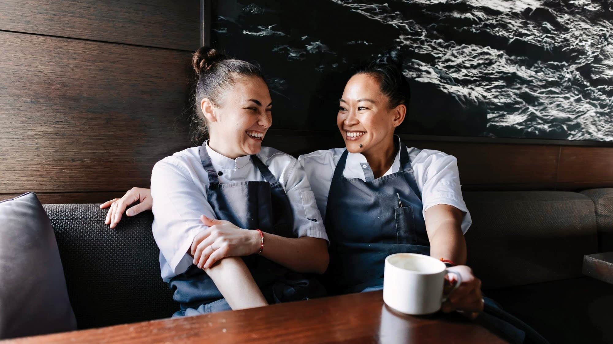 Two people in chef uniforms sit on a couch, smiling and looking at each other as if sharing a secret recipe. One holds a mug, their chemistry palpable. They are in a relaxed setting with a dark textured wall in the background, adding to the romantic ambiance.