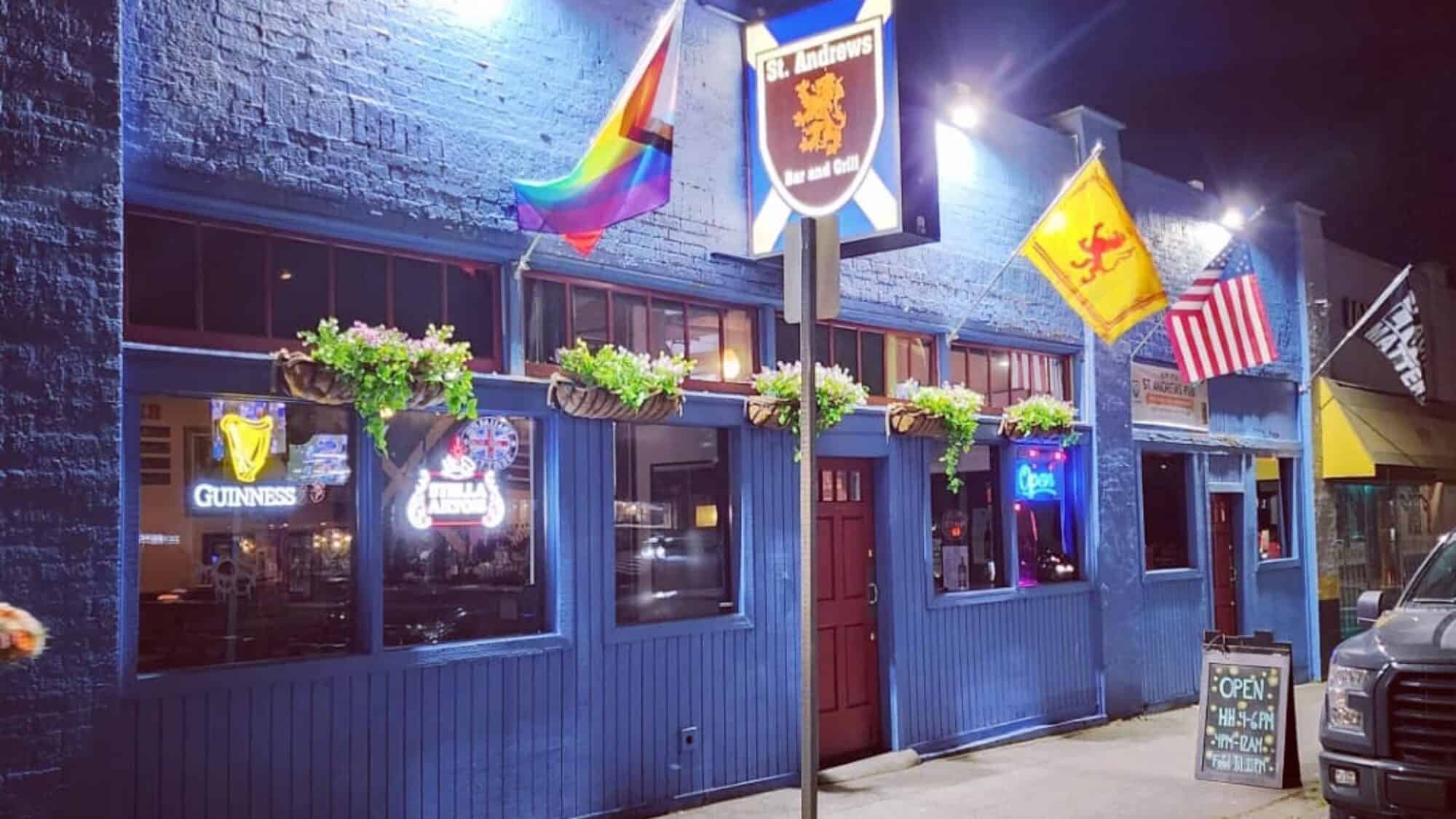 Blue brick pub exterior with three flags: rainbow, lion rampant, and American. Signs in windows include Guinness, Harp, and open. Flower boxes adorn the façade. Evening setting with street view.