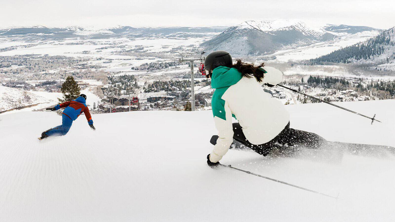 Two skiers in colorful jackets expertly descend a snowy slope, part of the best snow in Park City, overlooking a mountainous landscape and a distant town.