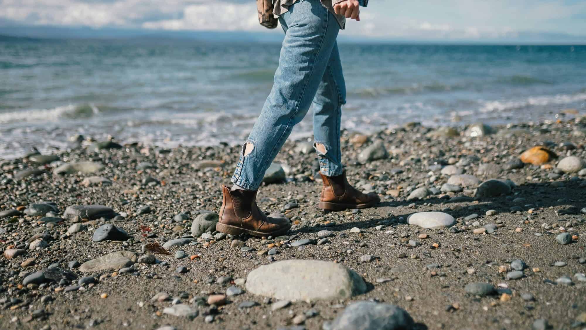 A person in jeans and boots takes a leisurely stroll along the rocky beach, near the water's edge.