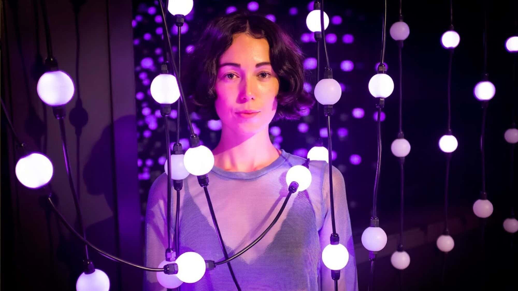 Person standing among hanging string lights with purple glow, in a dimly lit environment.