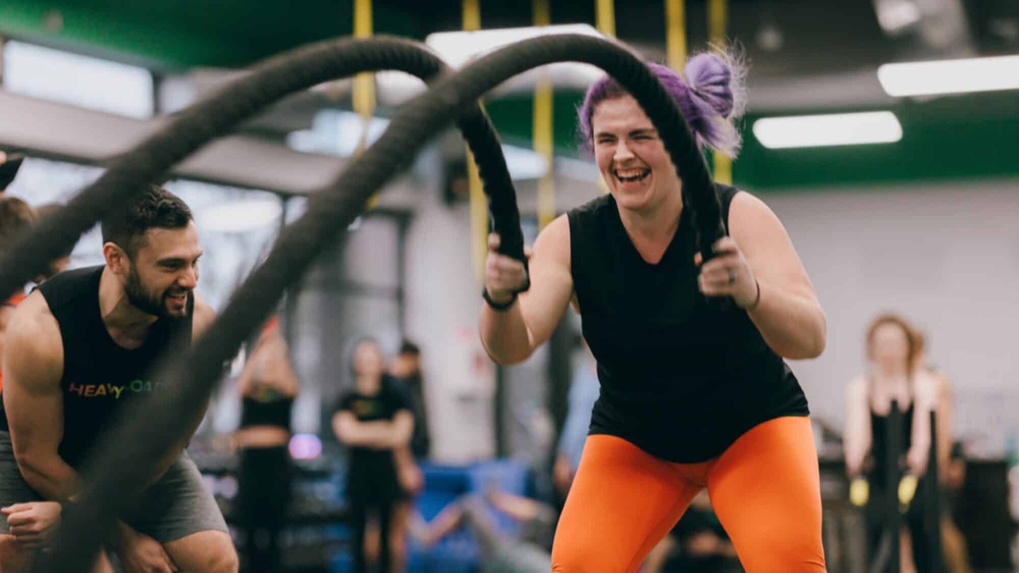 Person enthusiastically swinging battle ropes in a gym, wearing an orange outfit. An instructor is nearby offering encouragement.