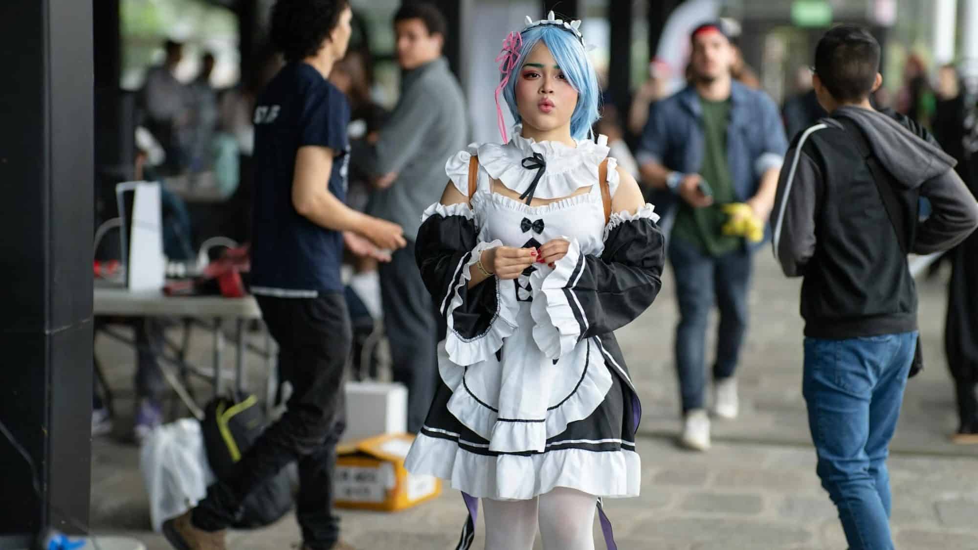 Person in a maid outfit with blue and pink hair walks through a crowded area, holding an object. People are in the background with tables and equipment around.