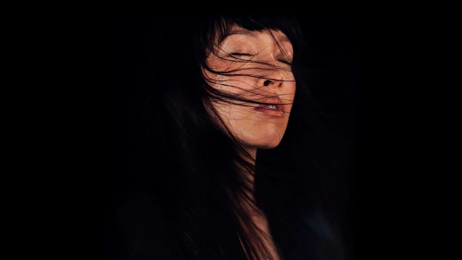 A woman with long dark hair, a true must-list for portrait lovers, closes her eyes as strands blow across her face against a black background. Captured on 04-24-2025.