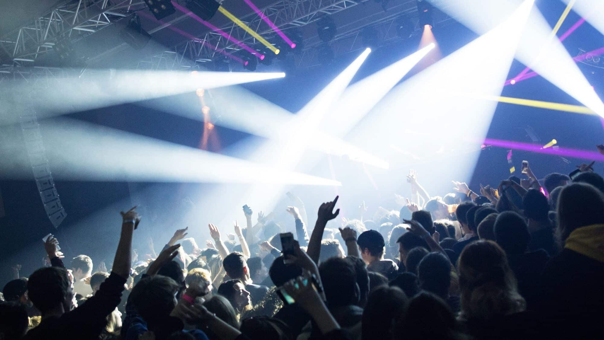 A crowd of people at a concert or music event, raising their hands and phones, with bright stage lights and colorful beams shining over them.