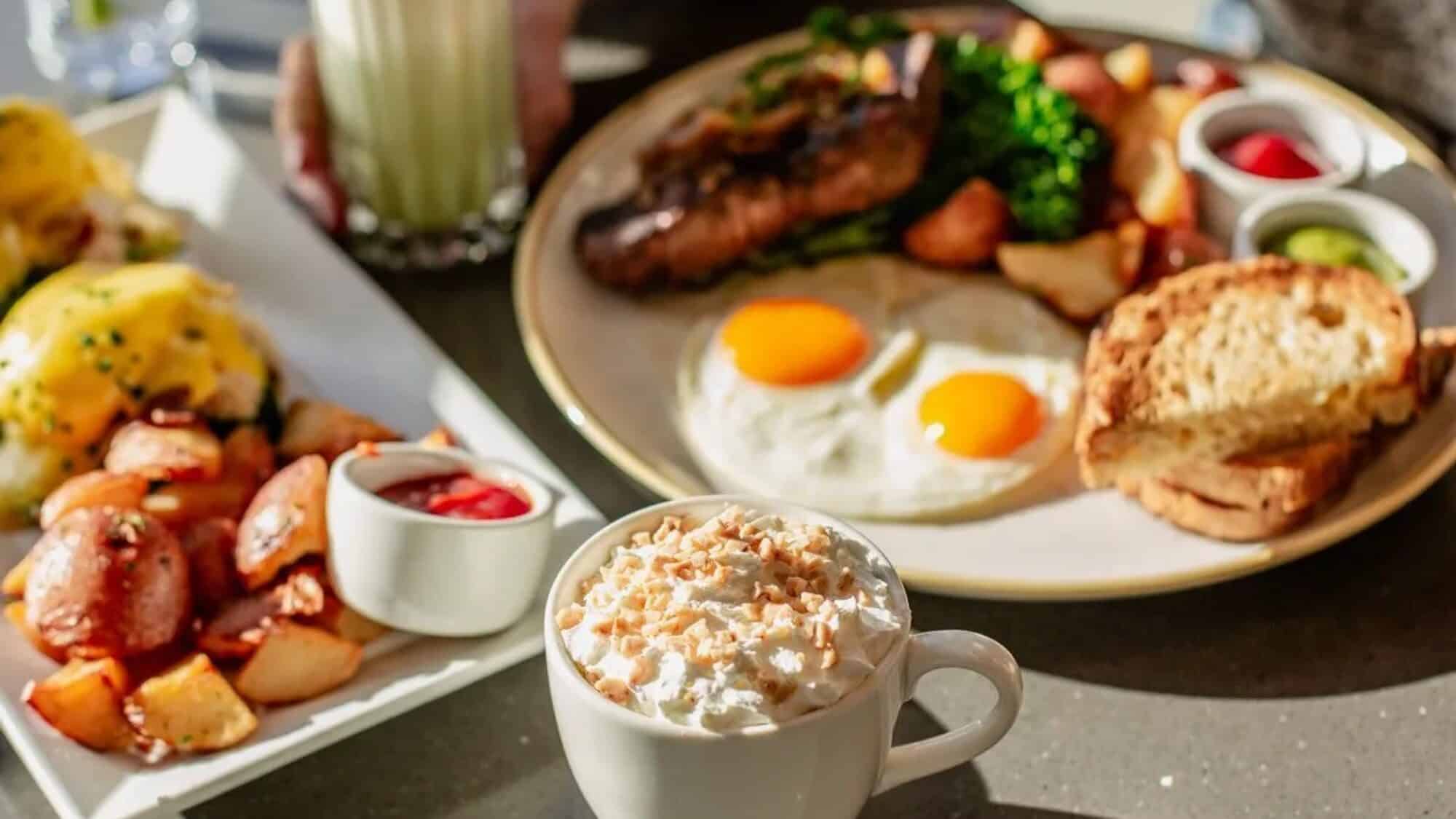 A table with a breakfast spread—including eggs, toast, sausage, roasted potatoes, broccoli, eggs Benedict, and a cup of whipped cream-topped coffee—awaits as guests scramble for a table.