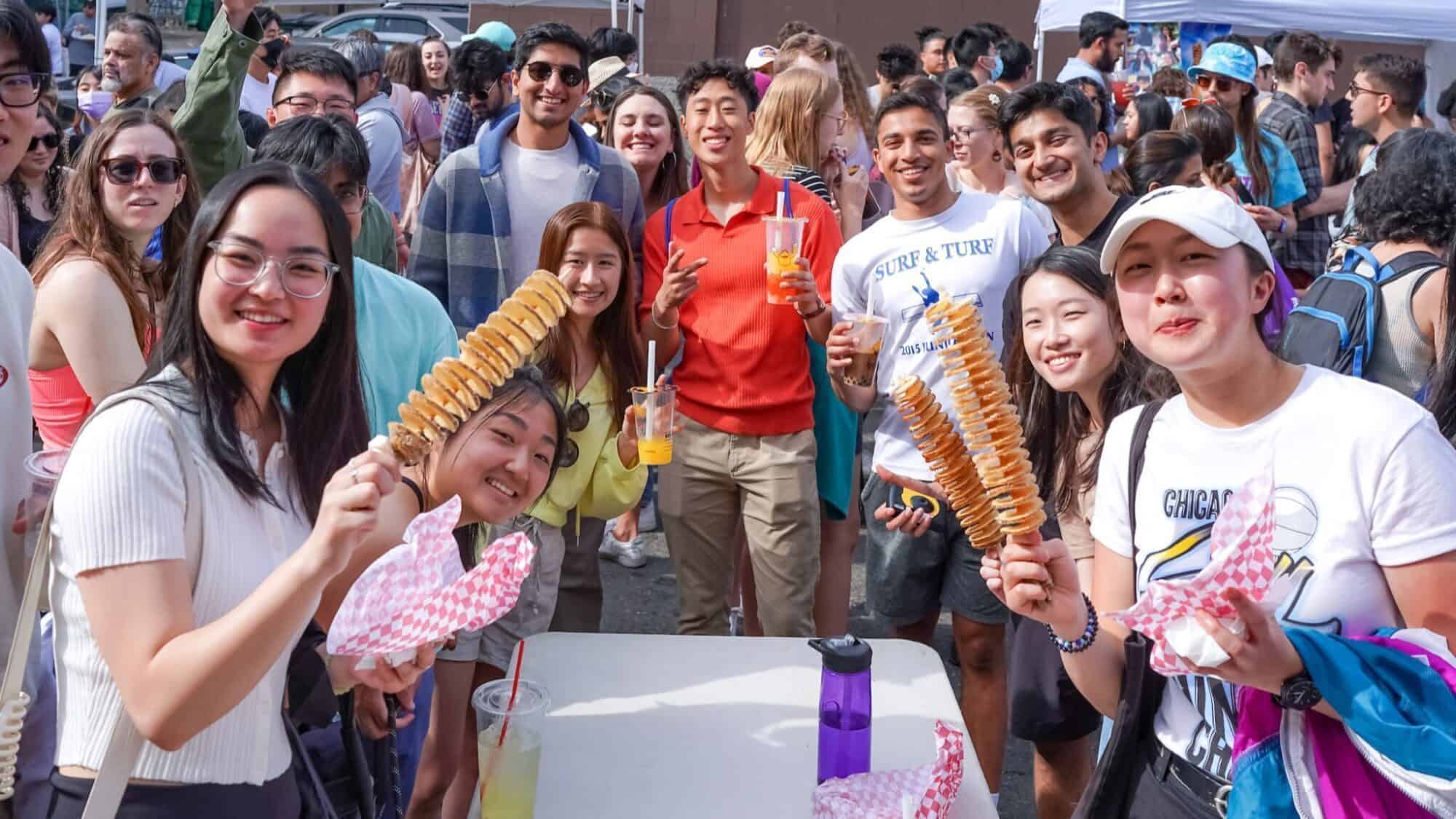 A group of young adults gathers around a table at an outdoor event, holding skewered spiral potatoes and drinks, smiling at the camera.