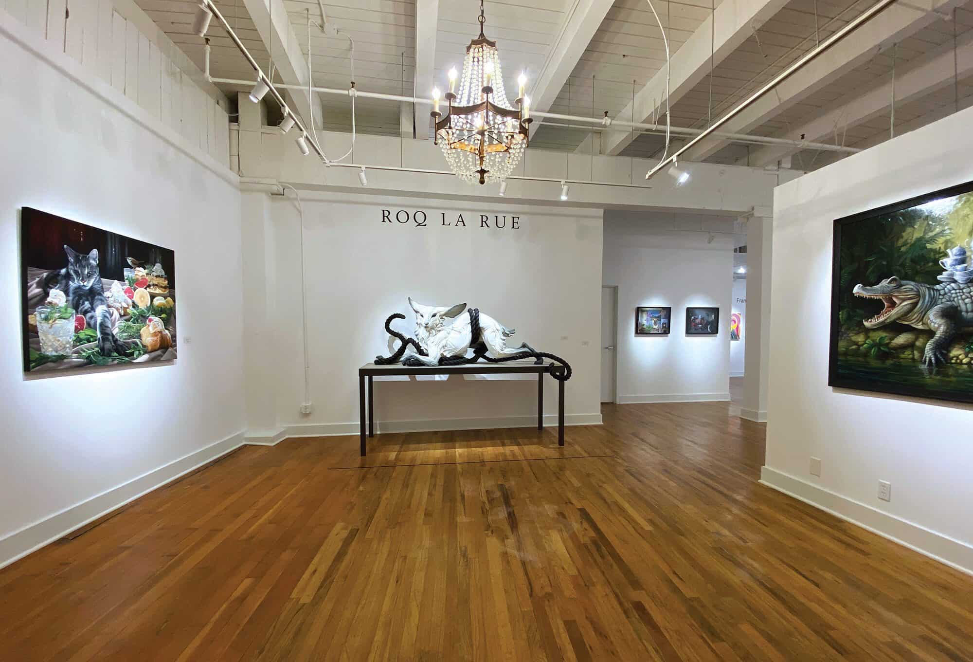 Art gallery interior with wooden floors, featuring paintings of animals on the walls and a sculpted white cat with tentacles, displayed centrally under a chandelier—evoking the unique aesthetic of gallerist Kirsten Anderson's Roq La Rue art space.