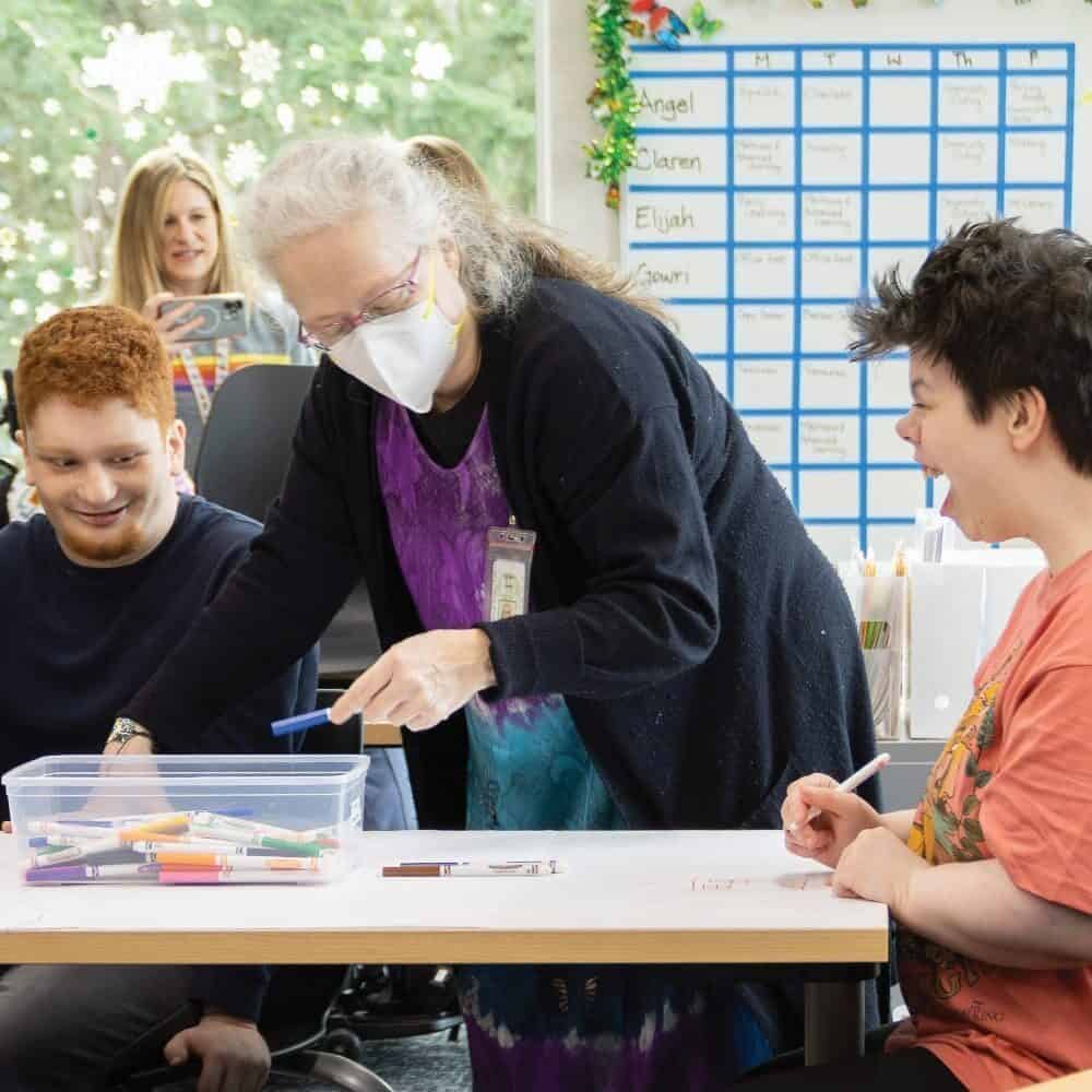 Adults from the Evergreen Transition Program, including students with disabilities, sit and stand around a table working on an art project with markers in a classroom setting.person