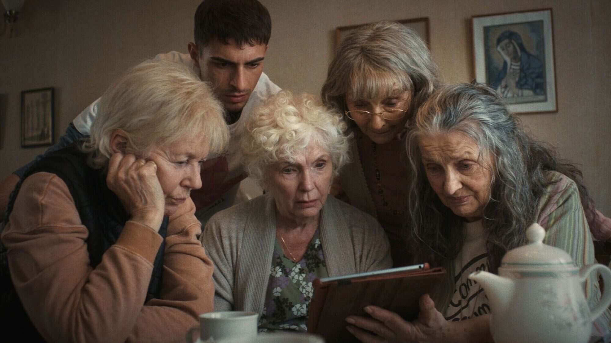 Five elderly women and a younger man closely look at a tablet together around a table in a cozy, softly lit room with framed art on the walls.