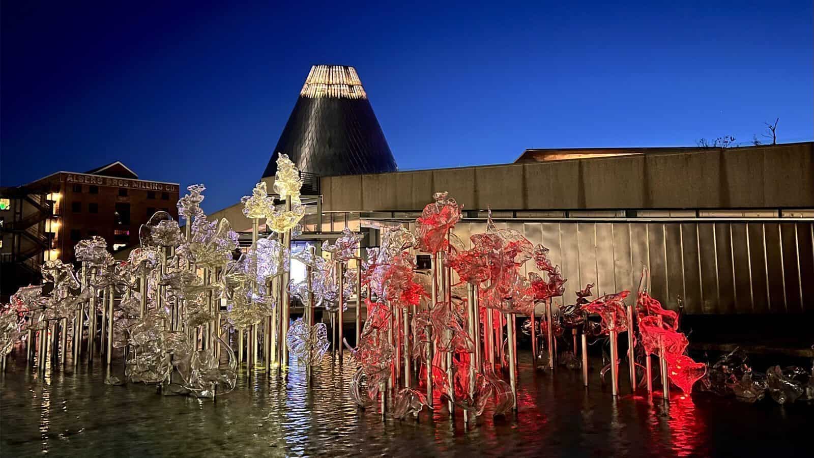 Glass sculptures on metal poles stand in shallow water, lit with white and red lights at dusk, capturing the essence of Heart of Glass: A Weekend in Tacoma, with a cone-shaped glass building glowing in the background.