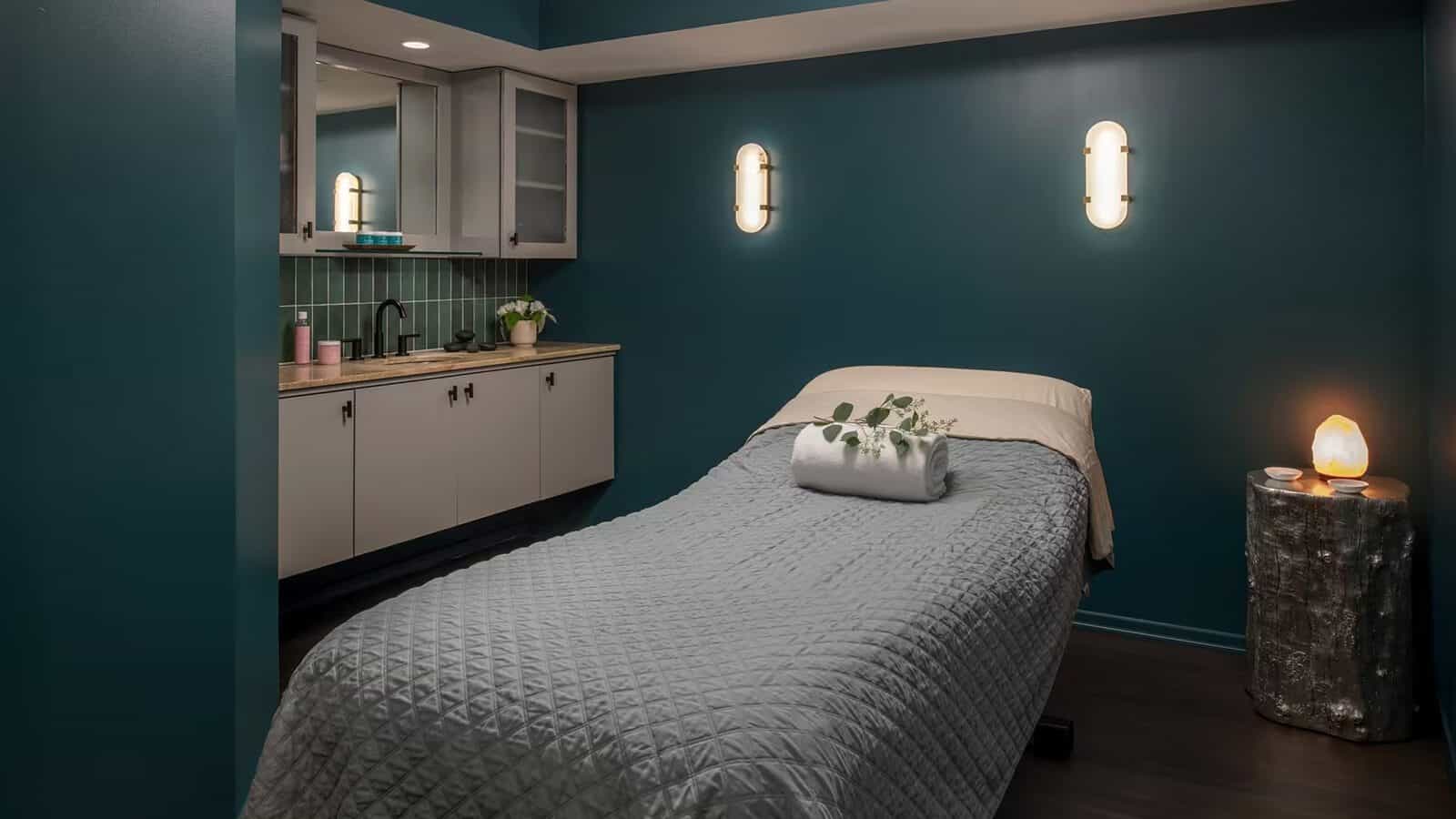 Treat Mom to some Emerald City Elegance this Mother’s Day in a tranquil massage room featuring a neatly made bed, rolled towel with leaves, soothing wall lights, a sink with cabinets, and a glowing salt lamp on a small table.