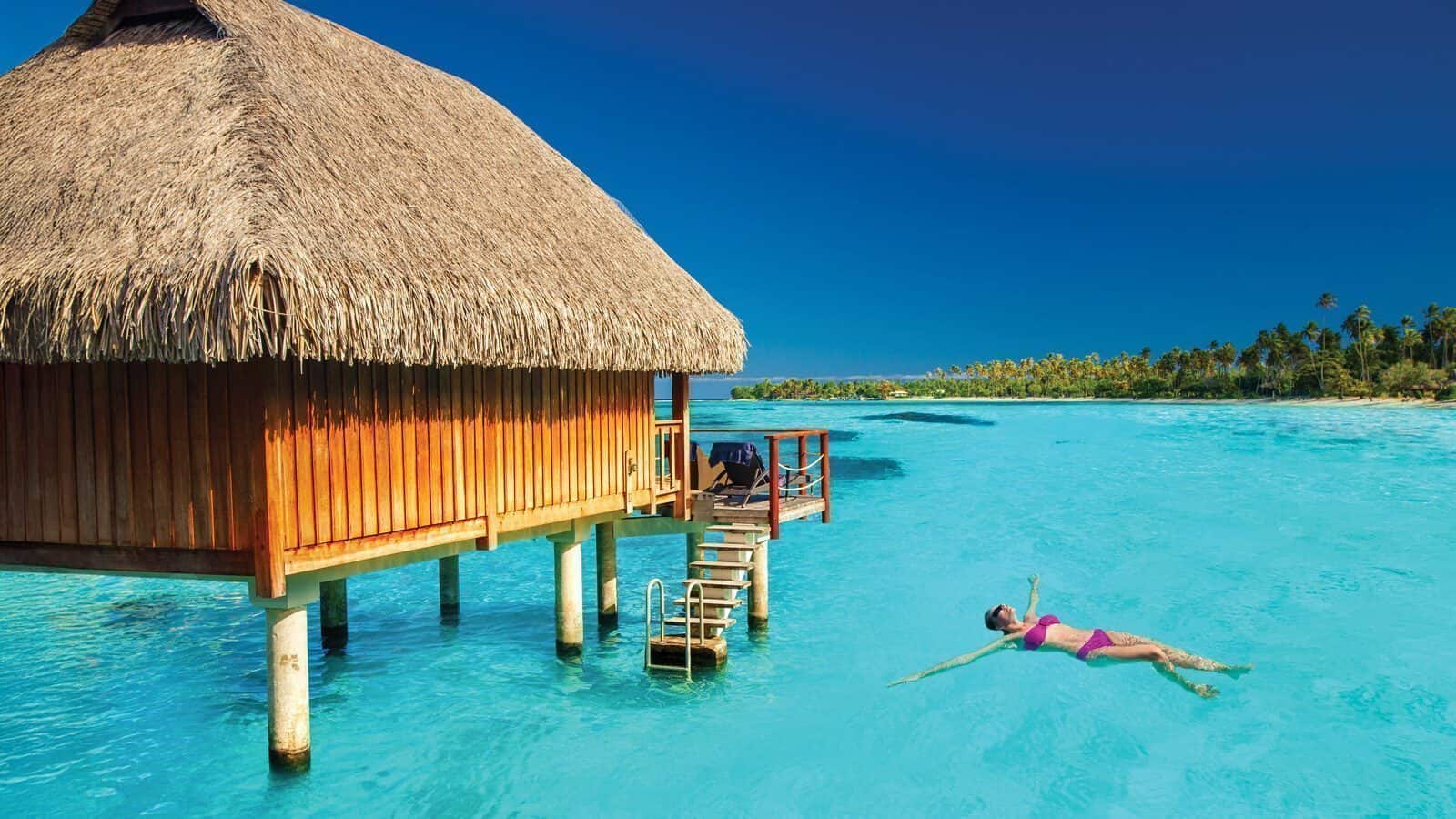 A person swims in clear turquoise water near a wooden overwater bungalow with a thatched roof under a bright blue Tahiti sky.
