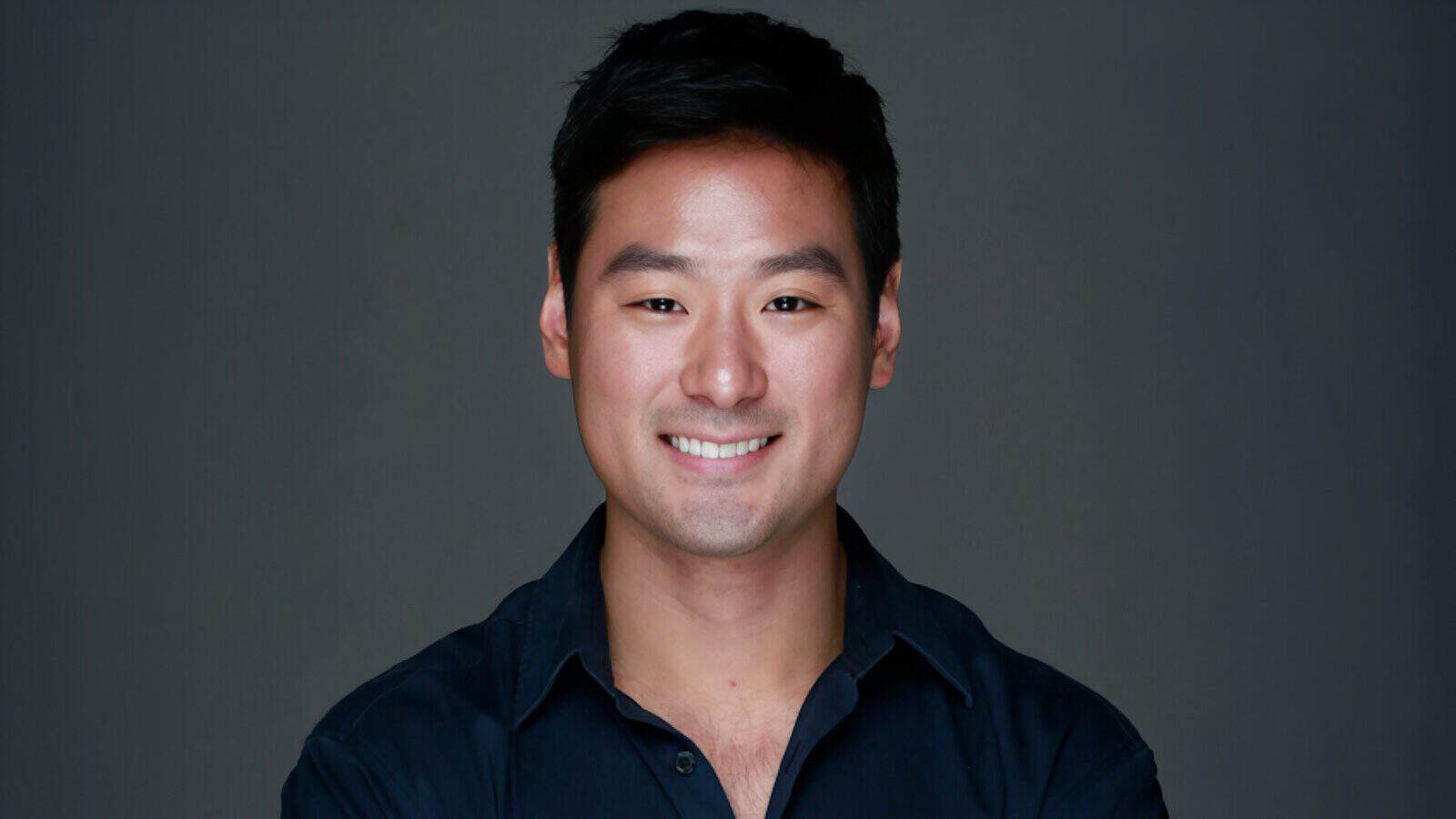 A man with short black hair and a dark shirt smiles at the camera against a plain dark background—Music Producer Kyu Lee talks Seattle, UW, and his Hollywood career.