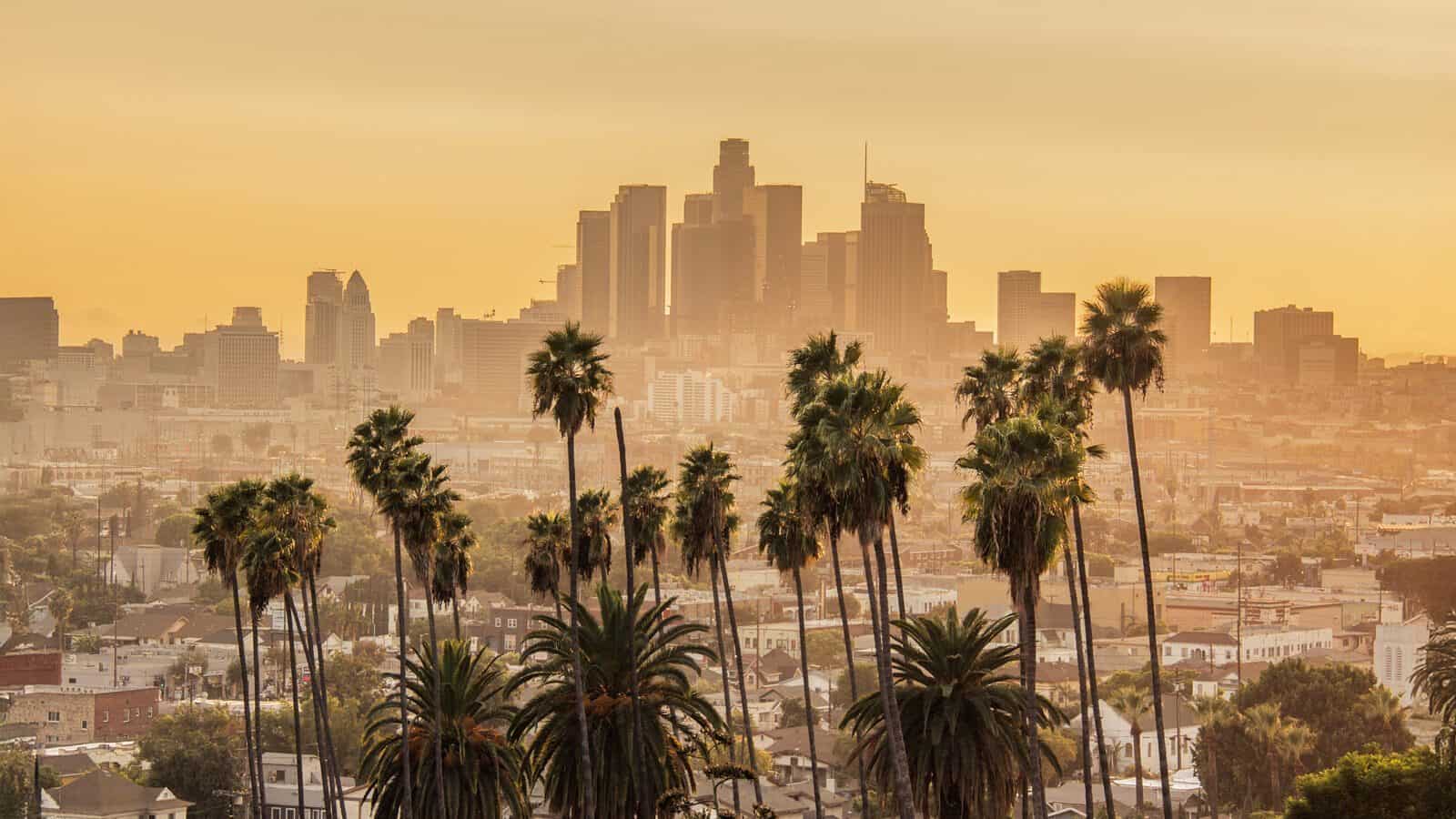 Downtown Los Angeles skyline at sunset, tall palm trees in the foreground and city buildings behind, as smoke fades, hope rises in L.A. beneath a golden sky.