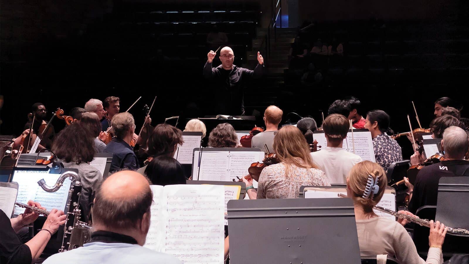 A conductor leads an orchestra during a rehearsal at the Shorewood Performing Arts Center, with musicians reading sheet music beneath dim lights as they prepare for the Mahler Fest 30th anniversary.