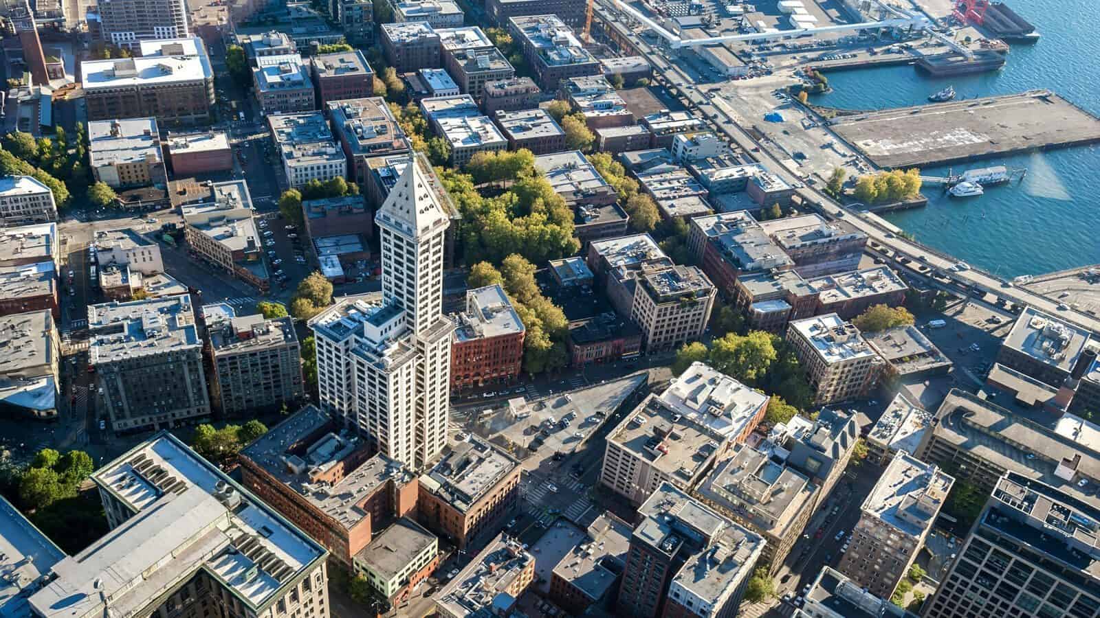 Aerial view of a cityscape featuring a tall, narrow tower surrounded by rectangular buildings, tree-lined streets, and a waterfront area with docks.