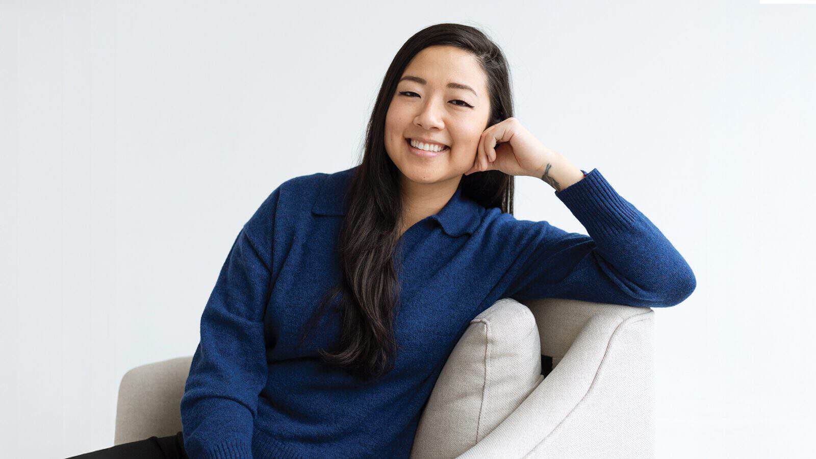 Joan Sung, a Korean American woman, with long dark hair and a blue sweater, sits on a light-colored chair, smiling with her head resting on her hand against a pale background.