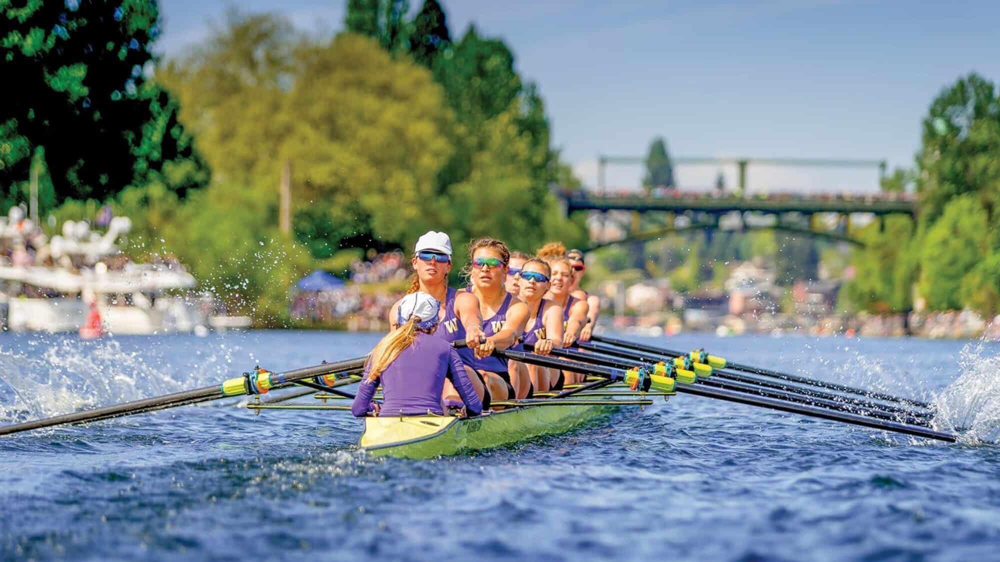 In May, a women's rowing team in matching uniforms and sunglasses rows a yellow boat at full speed on a river, with trees and a bridge in the background.