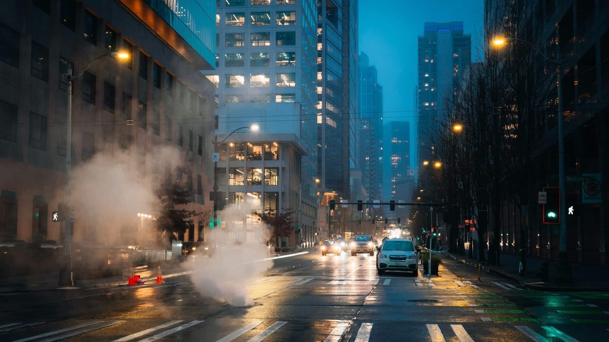 A city street at dusk with wet pavement, steam rising from a vent, cars driving, and tall buildings illuminated by streetlights and office lights.