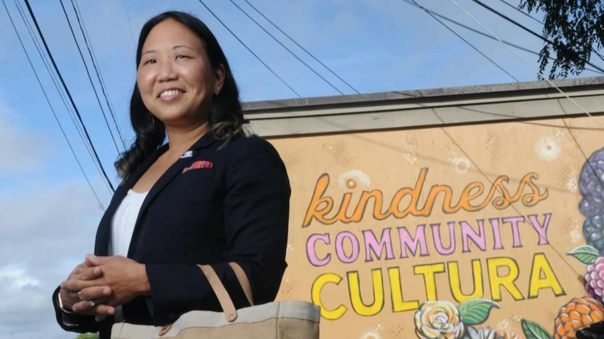A person stands in front of a mural with the words "kindness COMMUNITY CULTURA" and floral designs, holding a beige bag and smiling.