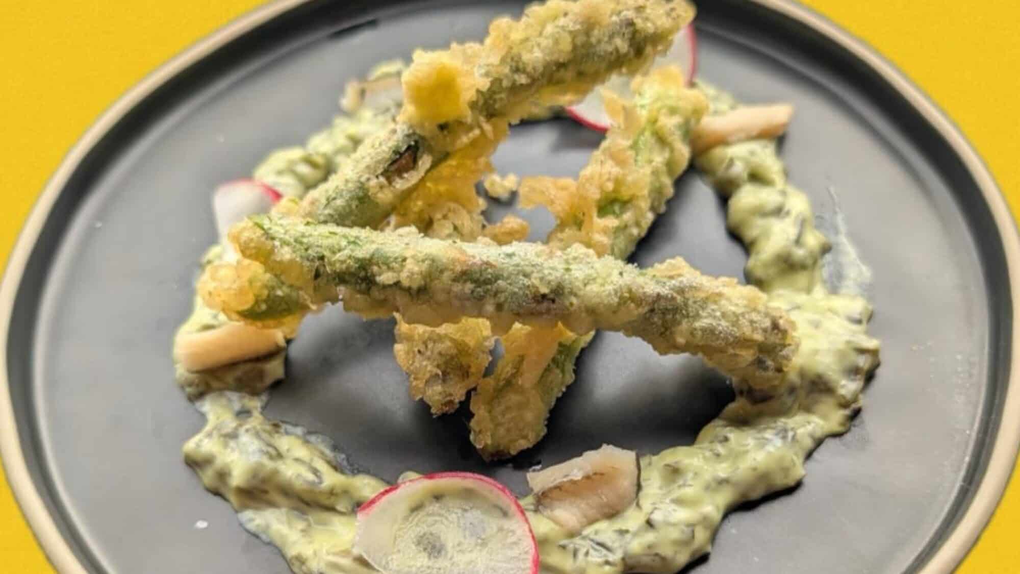 Three pieces of tempura asparagus are arranged on a black plate over a swirl of creamy green sauce, garnished with radish slices and small white vegetable pieces.