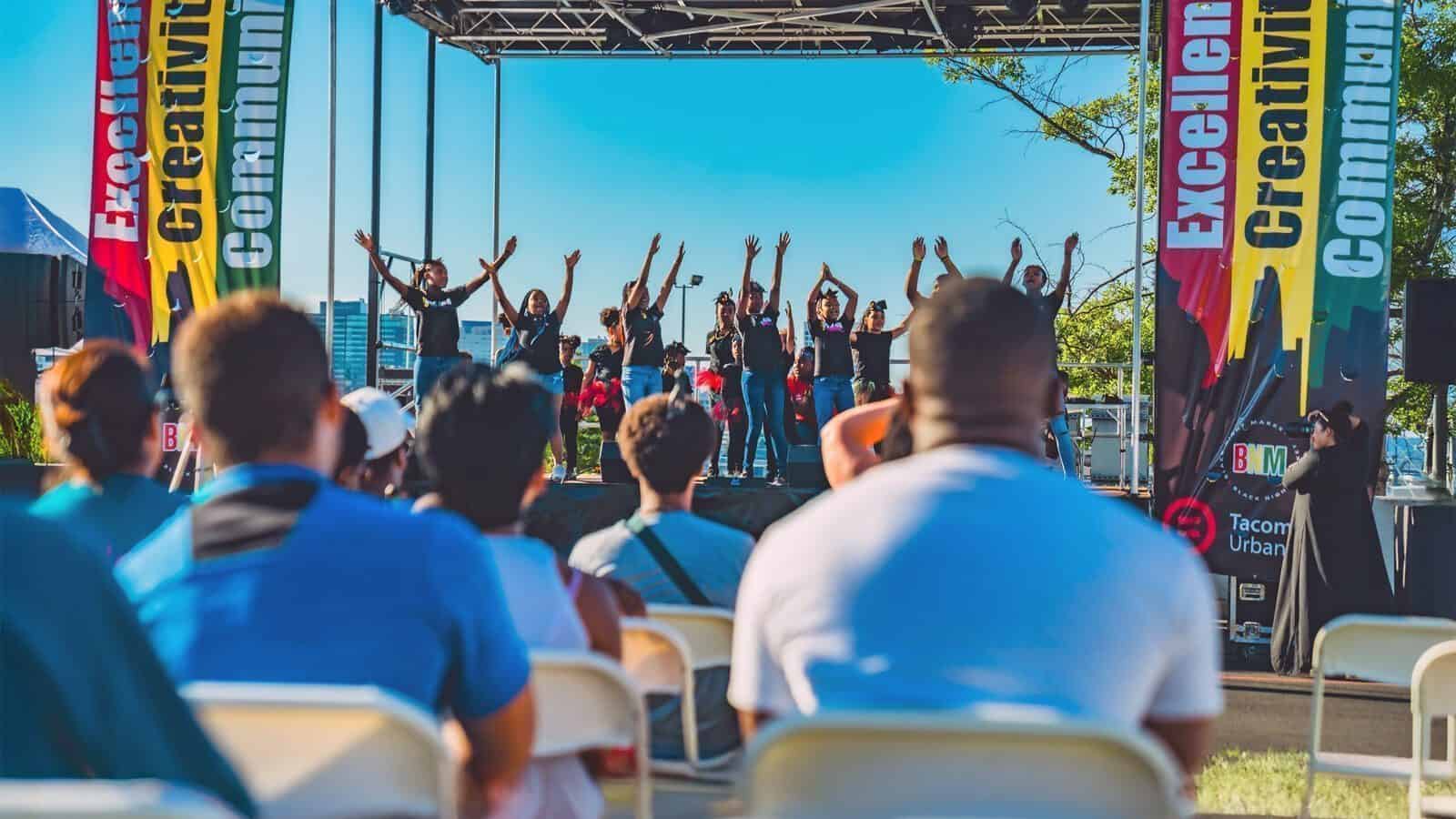 A group of performers dance on an outdoor stage as an audience watches; banners on stage read “Excellence” and “Creativity.”.