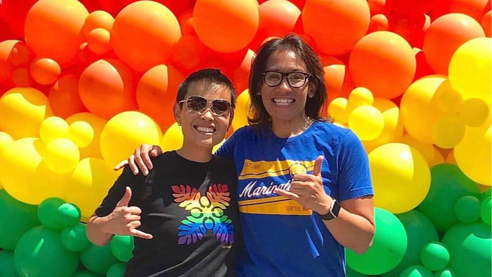 Two people smile and pose in front of a wall of colorful rainbow balloons, both making hand signs and wearing casual t-shirts.