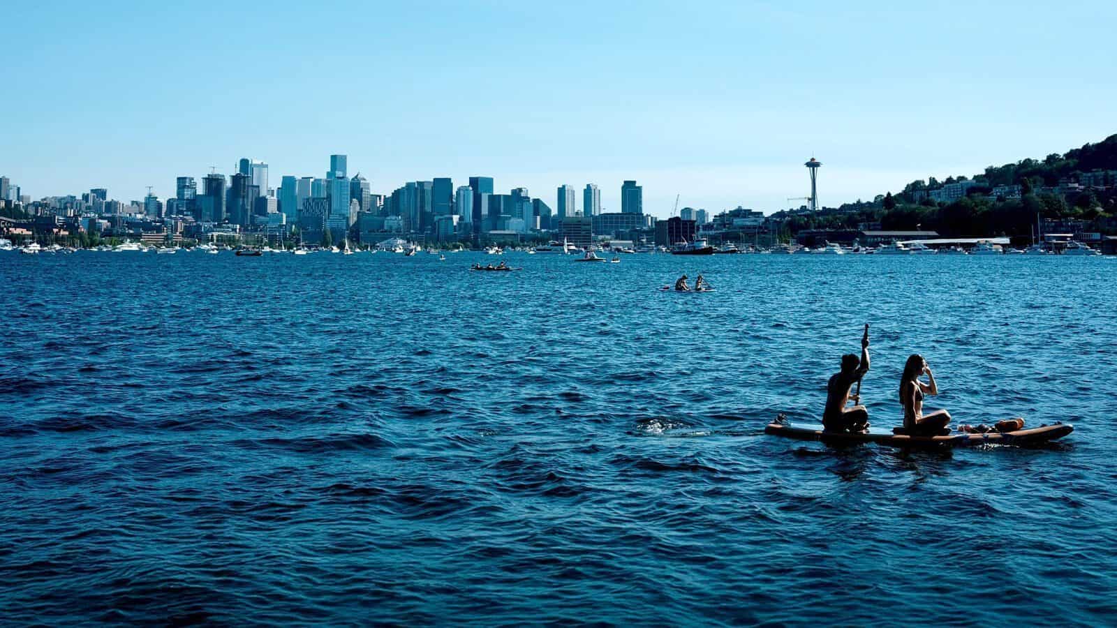 Two people paddleboard on a blue lake with the Seattle skyline, including the Space Needle, visible in the background under a clear sky.