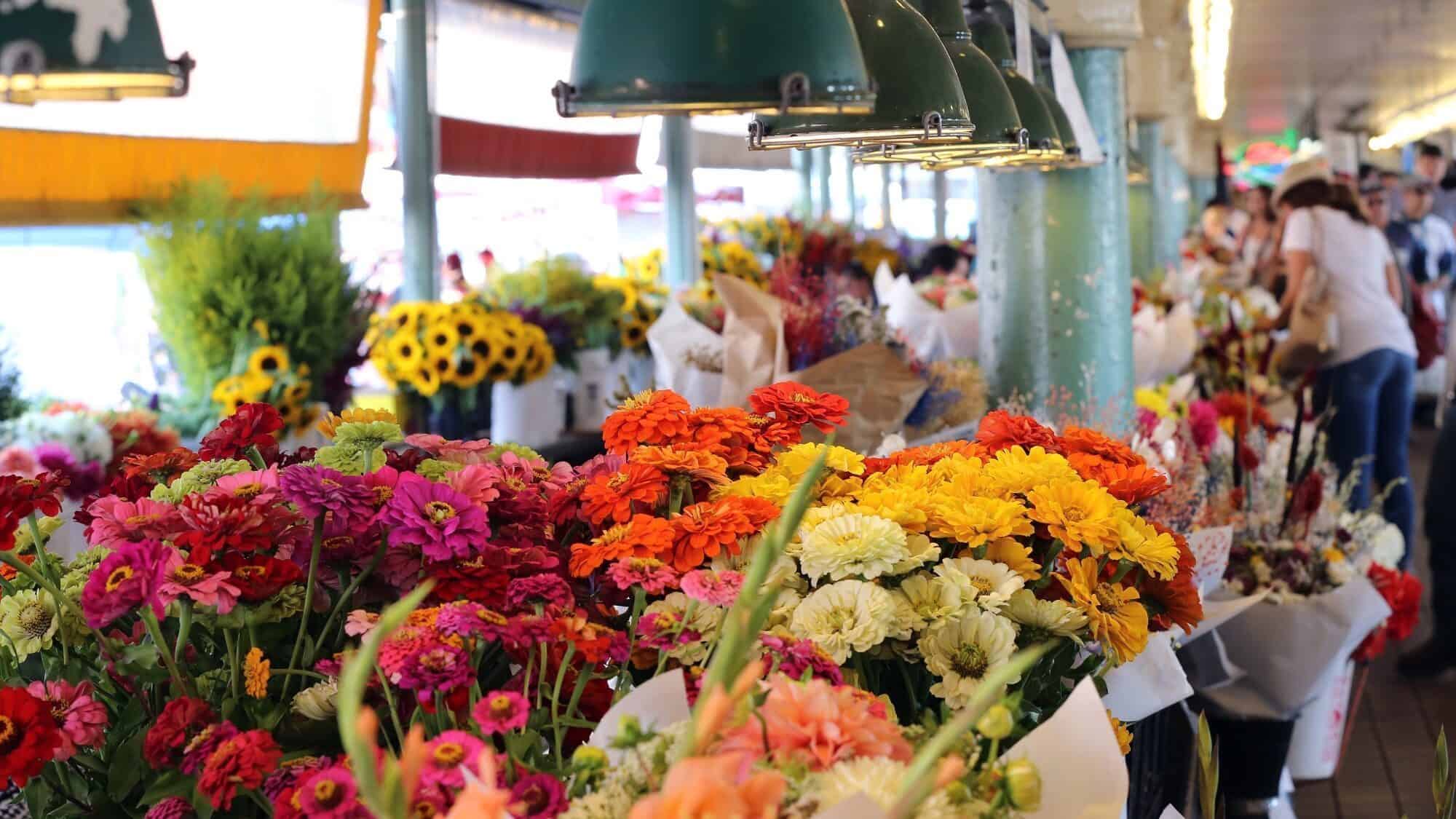 A market stall displays colorful bouquets of flowers under green hanging lights, with customers browsing in the background.