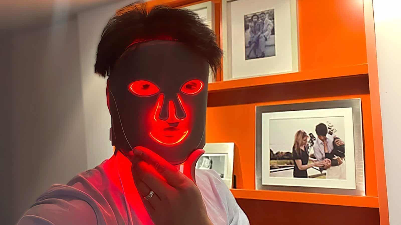 Person wearing an illuminated LED facial mask stands in front of an orange wall with framed photographs, highlighting their commitment to a skincare routine aimed at looking and feeling younger.