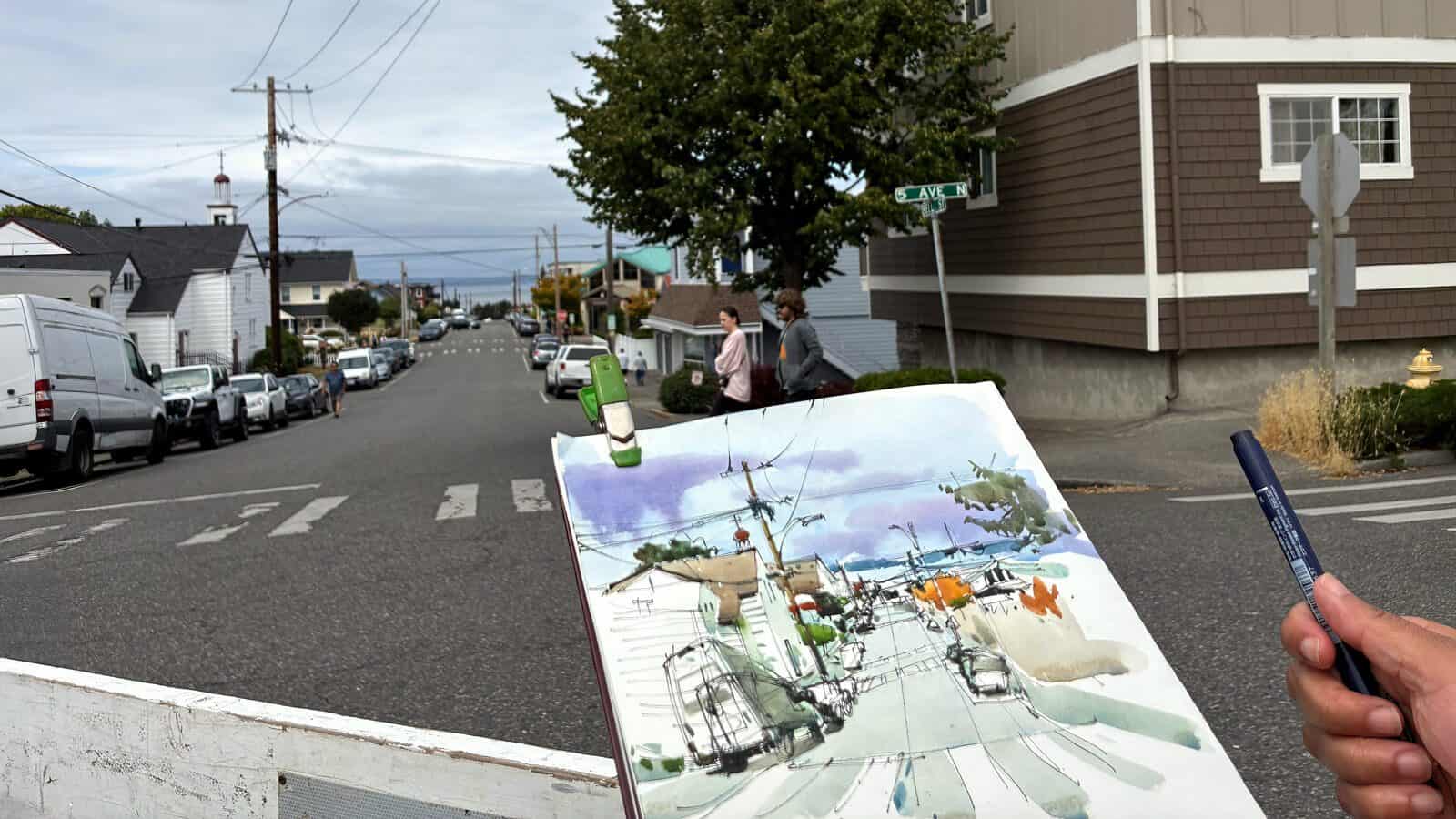 A person engaged in the Art of Looking holds a sketchpad and pen, painting a street scene that matches the neighborhood visible in the background.