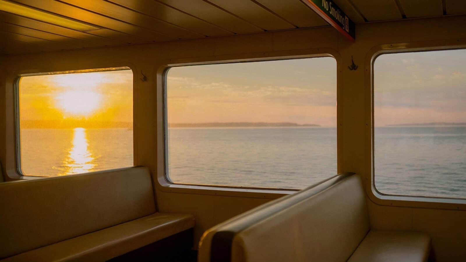 Empty ferry seats and large windows overlook a calm body of water at sunset, the summer light casting golden reflections on the waves.