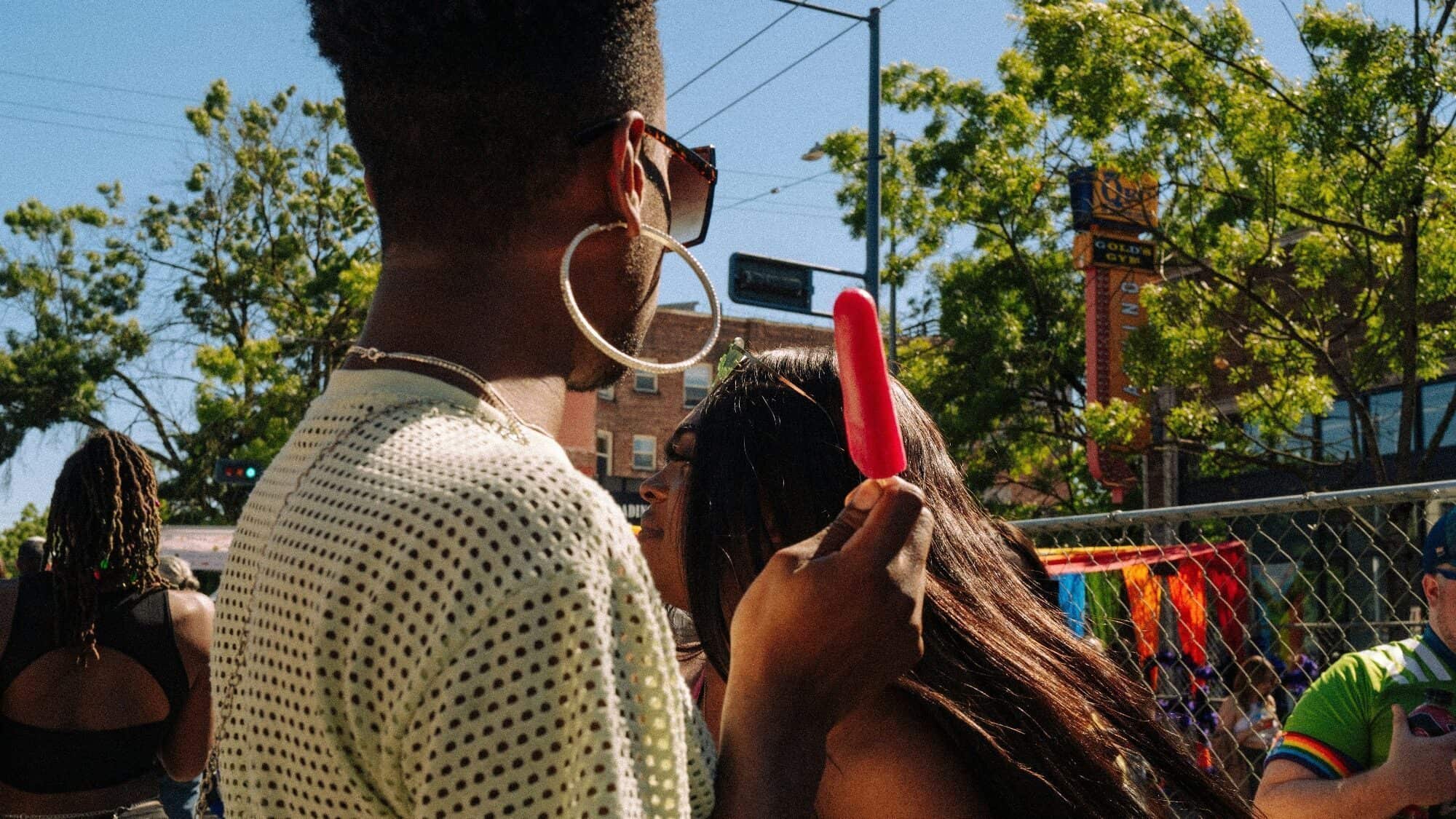A person wearing sunglasses and large hoop earrings holds a red popsicle at an outdoor event, capturing The Pulse of summer with Bring the Heat vibes; other people and trees are visible in the background.