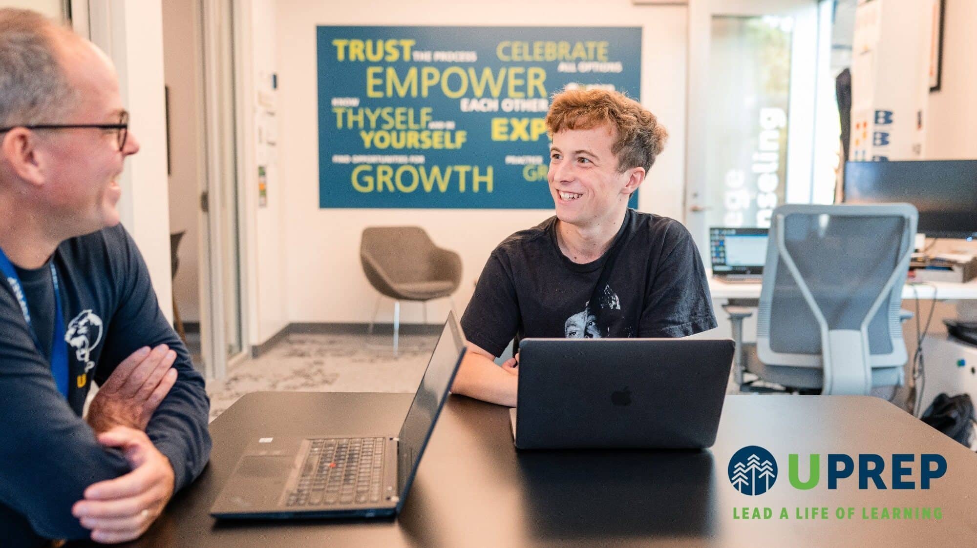 Two people sit at a table with laptops, discussing college admission in a modern office space adorned with motivational words and the UPrep private school logo in the foreground.