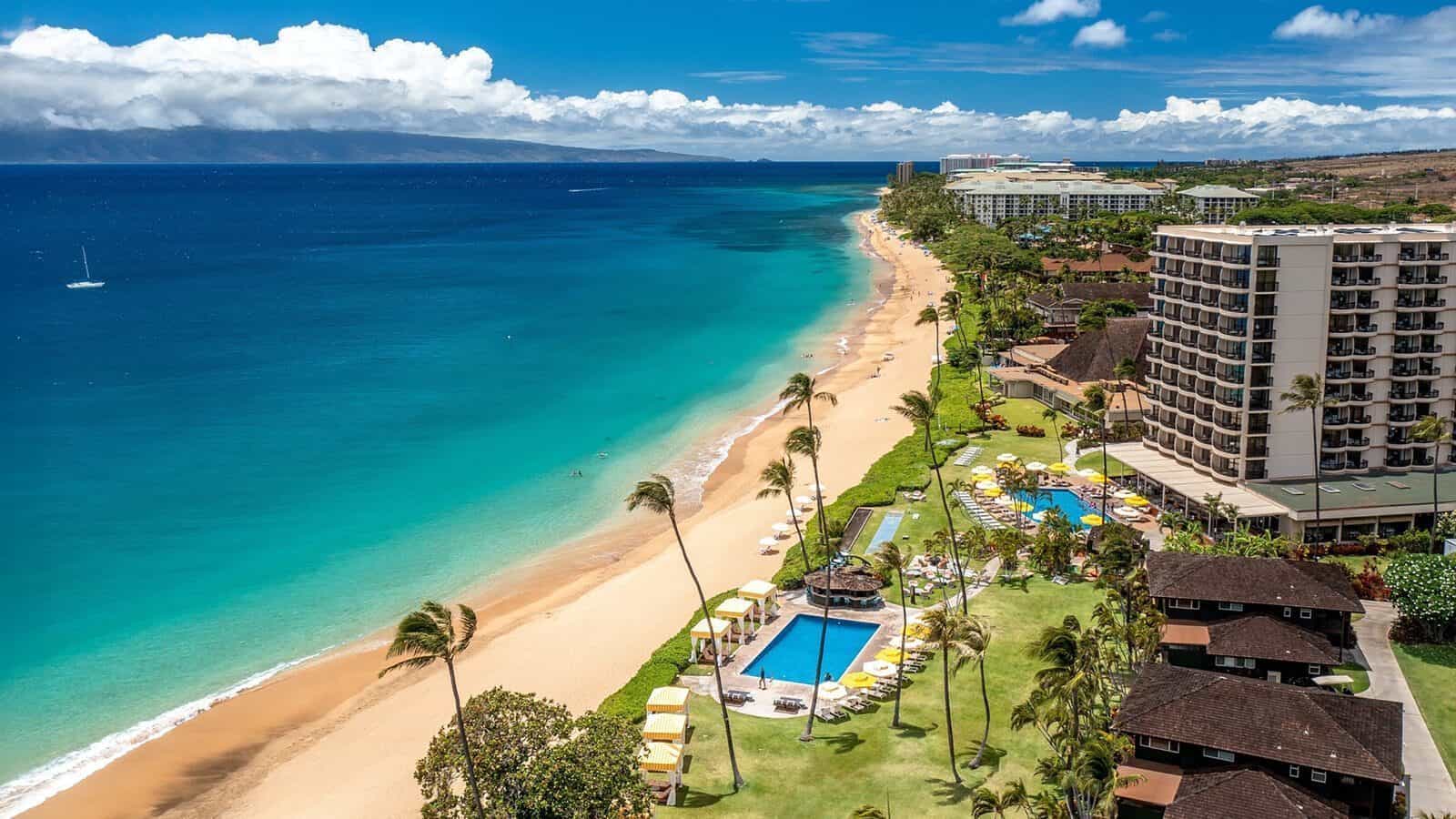 Aerial view of a tropical Lahaina beachfront resort with swimming pools, palm trees, sandy beach, and clear blue ocean under a partly cloudy sky—Resilience by the Sea.