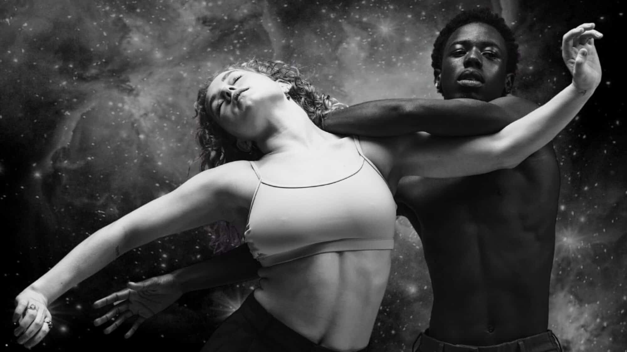 Two dancers, one male and one female, perform an expressive pose against a cosmic, star-filled background in black and white.