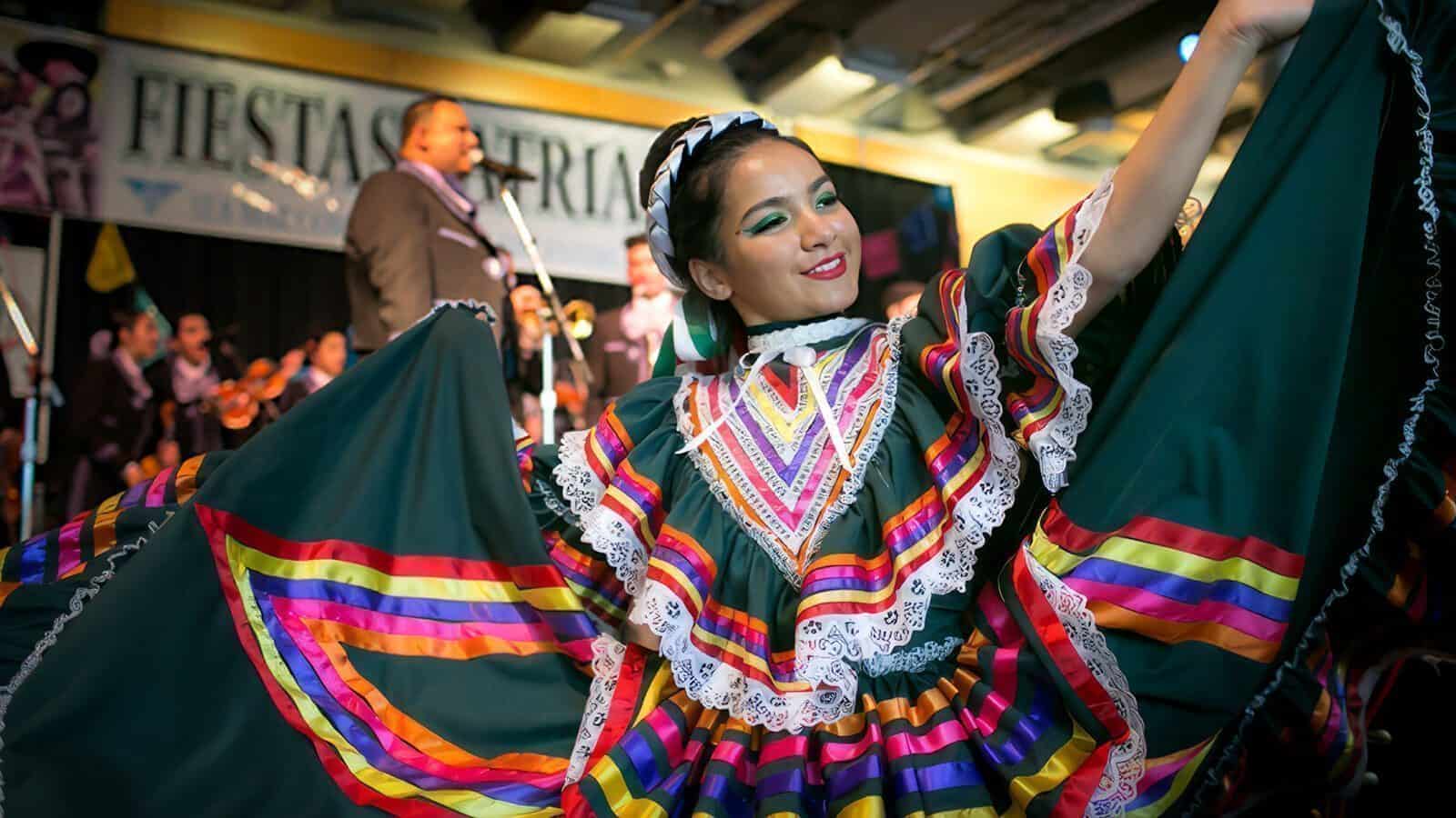 A dancer in a colorful traditional dress performs on stage at a Seattle cultural event, celebrating Hispanic Heritage Month, with musicians and a “Fiestas Patrias” banner in the background.