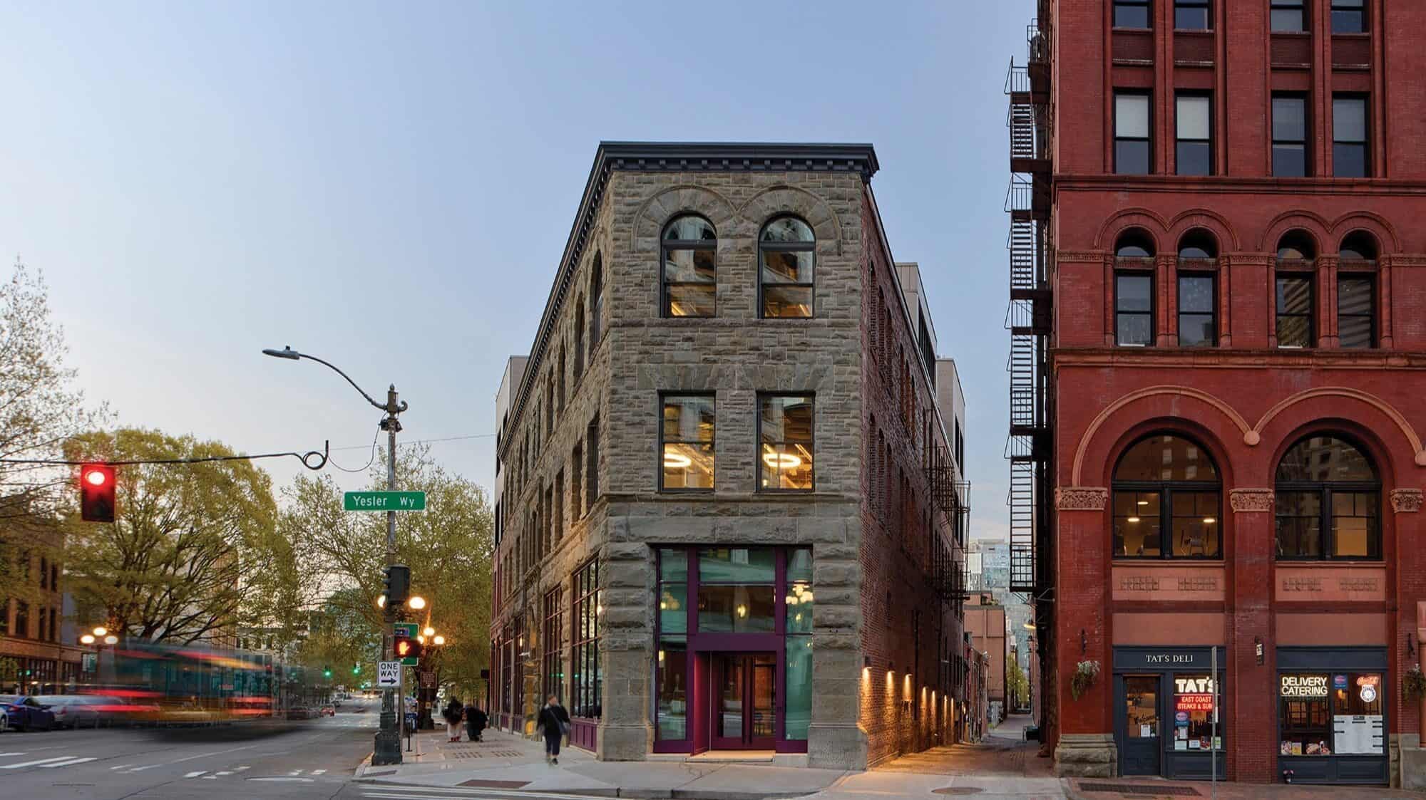 A stone and glass three-story building stands at a Pioneer Square street corner in Seattle, next to a taller red brick building, with trees and street signs visible.
