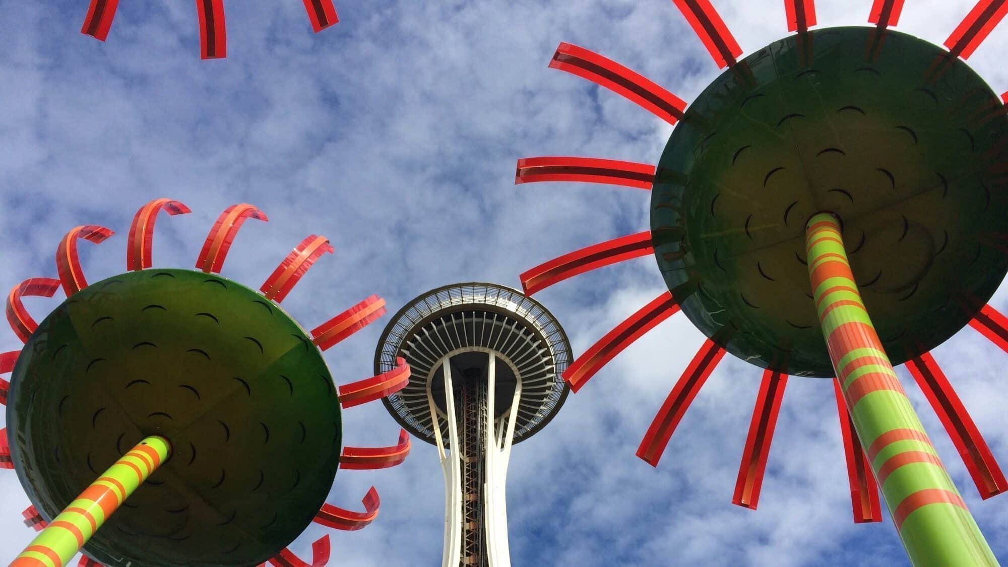 View looking up at the Seattle Space Needle framed by the colorful, striped Sonic Bloom art installation against a blue sky with clouds.