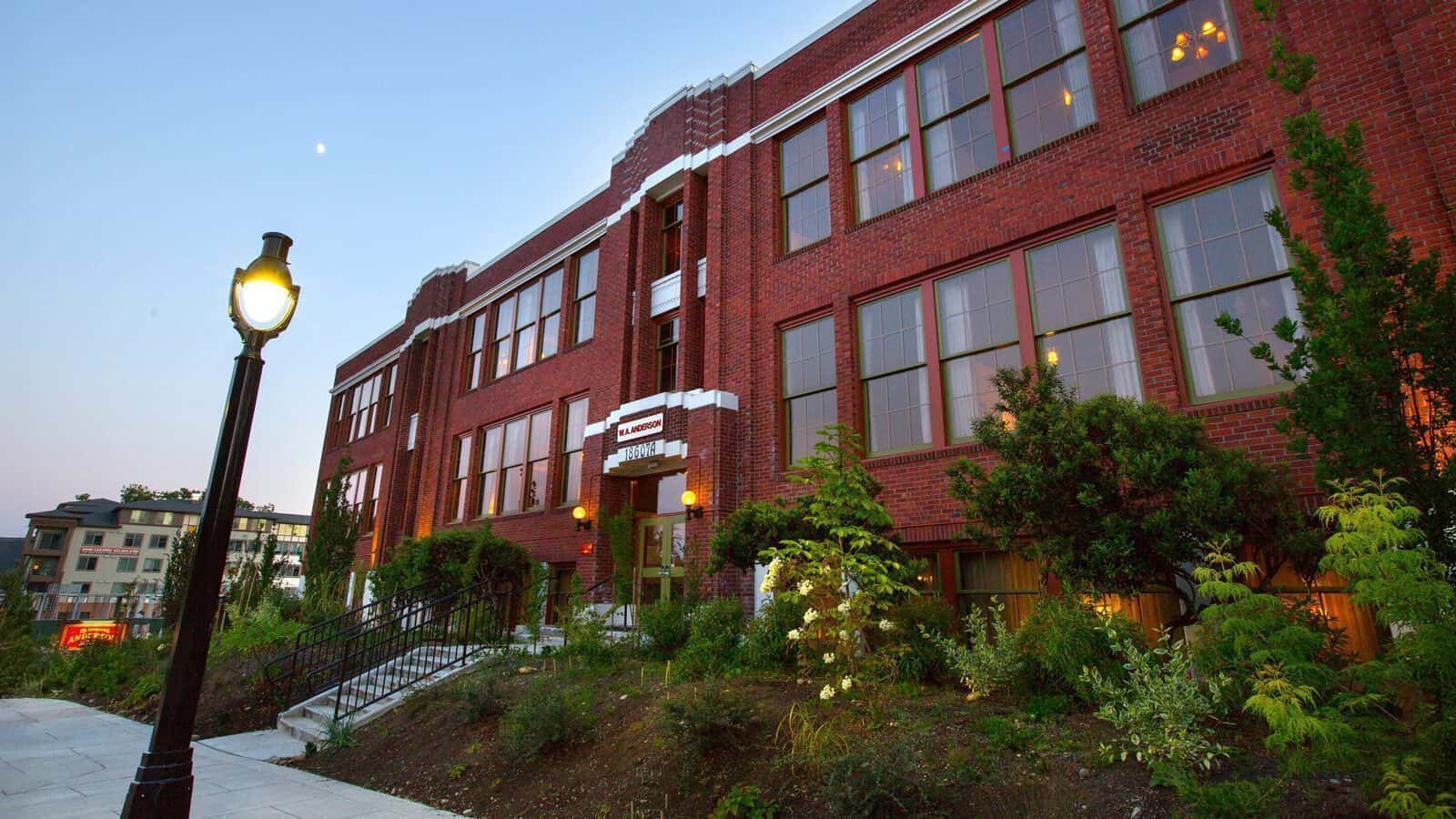 The Anderson School is a three-story red brick building with large windows, surrounded by shrubs and trees, next to a lit streetlamp and a concrete sidewalk, reflecting a commitment to preservation.