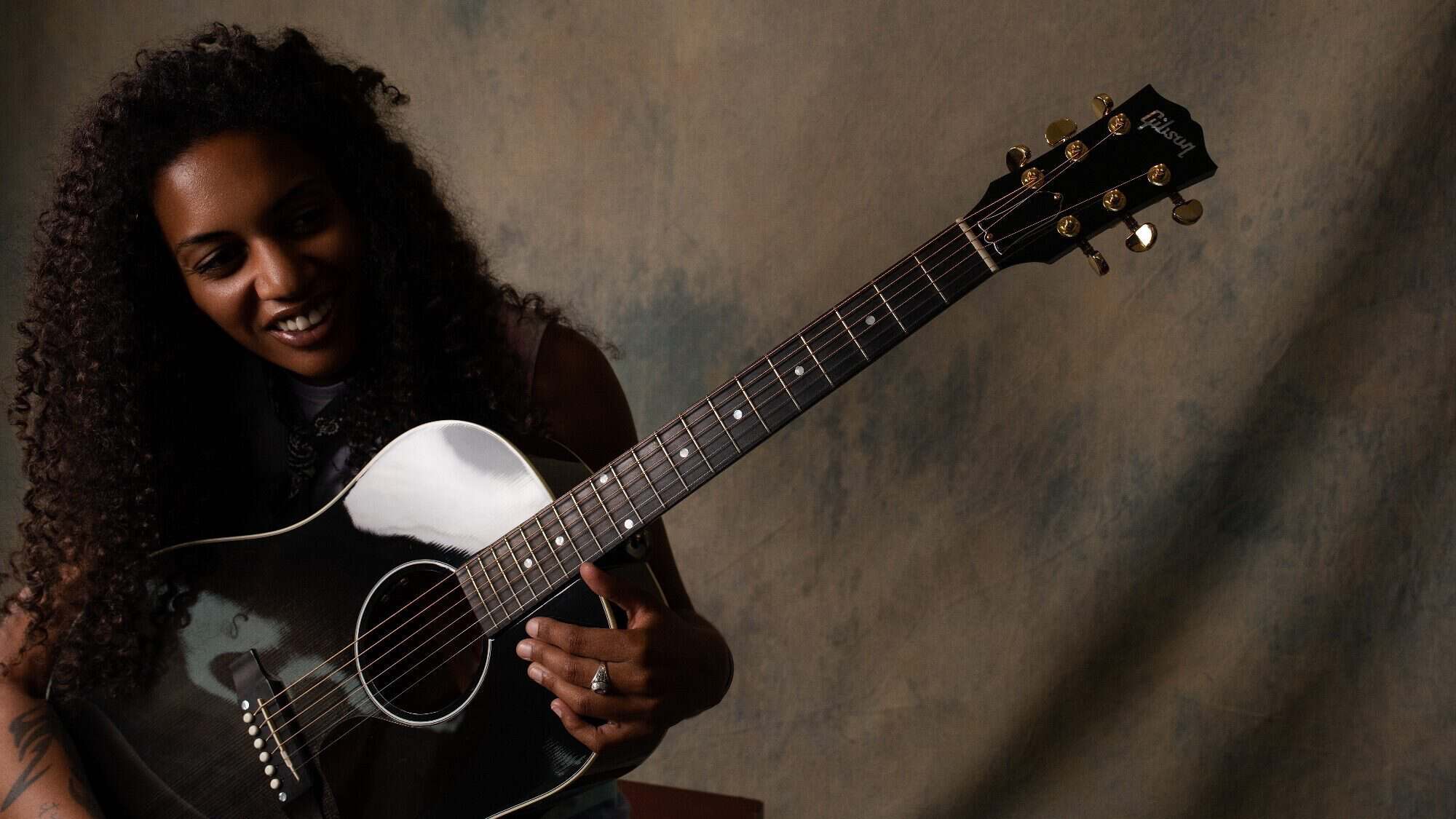 A person with long curly hair smiles while holding and playing a black acoustic guitar against a neutral background.