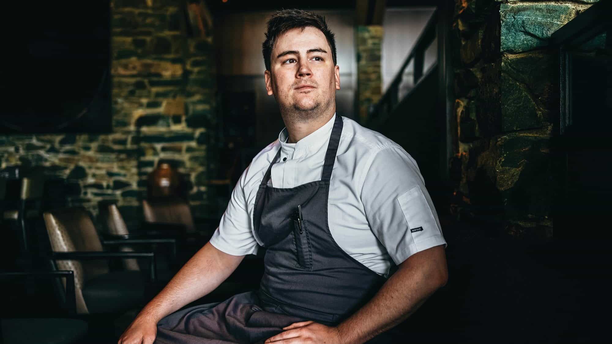 A chef in uniform sits on a chair in a rustic restaurant with stone walls, embodying the spirit of Hometown Harvest, looking off to the side with a neutral expression.