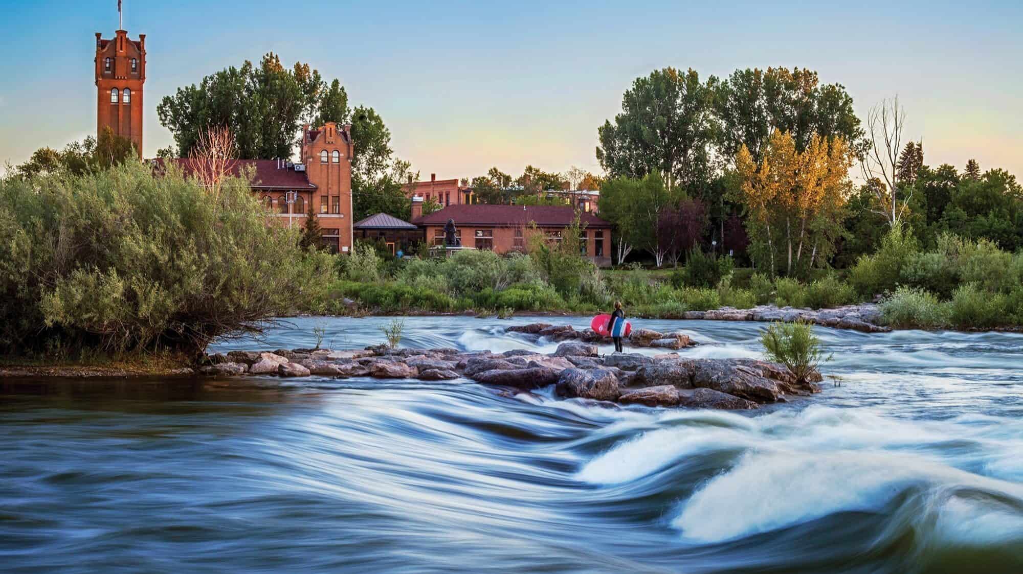 A person sits on rocks by a flowing river with historic brick buildings and trees in the background during sunset, capturing the charm of small towns and the scenic beauty that defines Montana tourism.