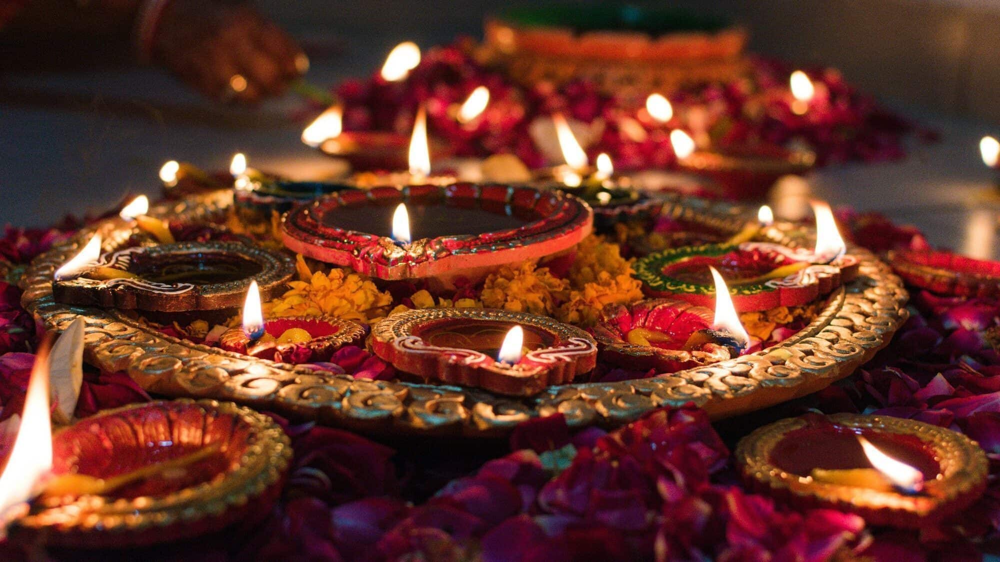 A decorative arrangement of lit clay oil lamps surrounded by flower petals, with a hand visible in the background, likely during a festive celebration.
