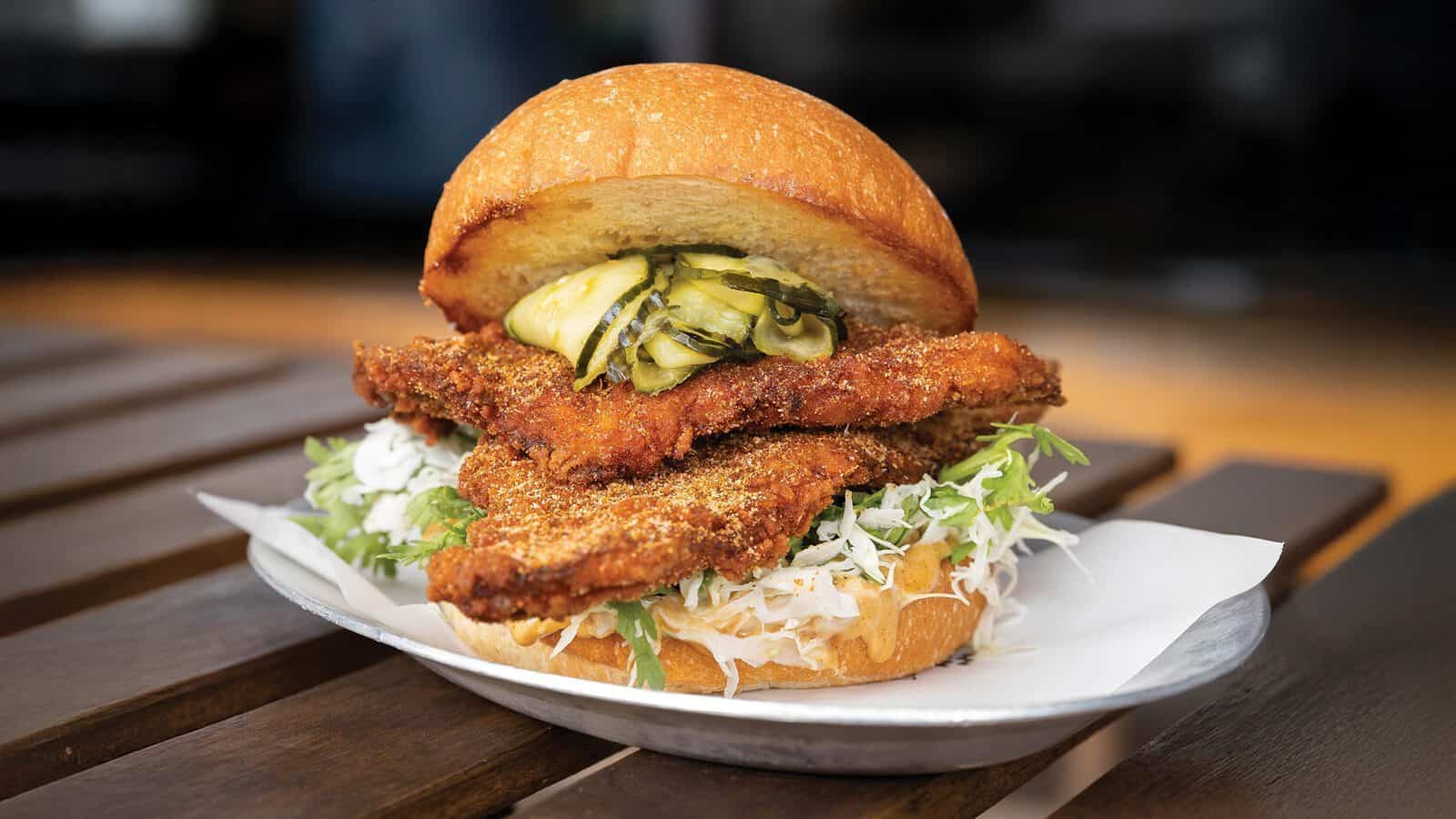 A Local Tide fried chicken sandwich with shredded lettuce, pickles, and a toasted bun is served on a plate lined with paper.
