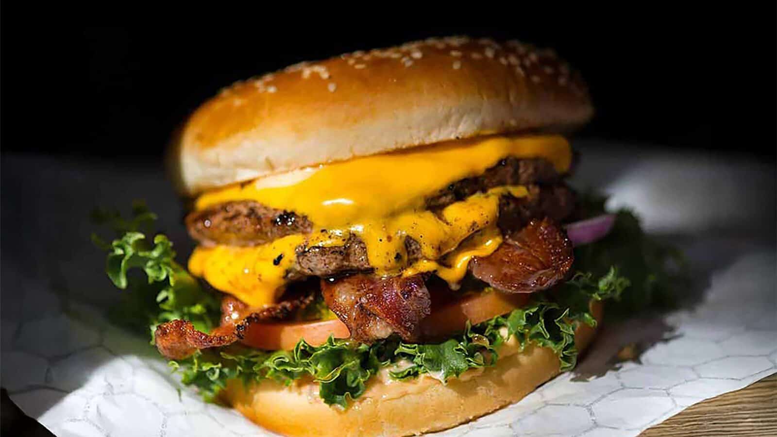 A Red Mill Burgers classic: a cheeseburger with juicy meat and melty cheese, lettuce, tomato, bacon, red onion, and two beef patties on a sesame seed bun. Enjoy it with crispy fries for the perfect meal.